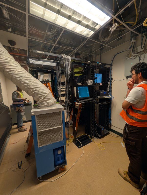 Cramped telecom room mid-teardown with a portable AC unit and disordered racks